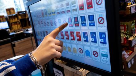 Eine Frau tippt an der Kasse eines Supermarktes mit dem Finger auf das Display von einem Zigarettenautomaten. 
