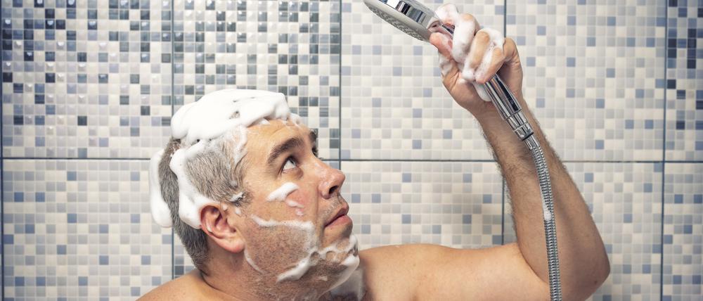 Ein Mann steht mit Shampoo im Haar unter der Dusche und blickt unsicher auf den Duschkopf in seiner Hand.