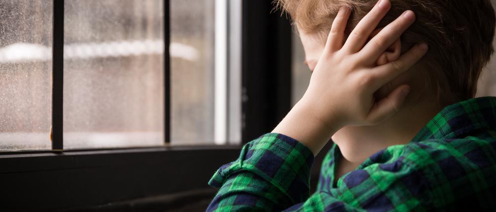 Ein kleiner Junge sitzt traurig vor dem Fenster und hält sich die Ohren zu.