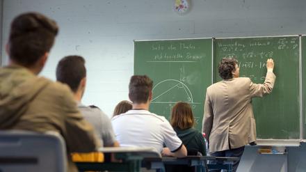 Ein Mathematiklehrer steht mit dem Rücken zur Klasse und schreibt mit Kreide Mathematikaufgaben an eine Tafel.